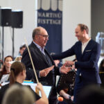 Beni Thurnheer spricht mit dem Publikum, im Hintergrund ist das Orchester und der Dirigent zu sehen.