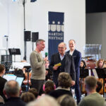 Alex Frei und Beni Thurnheer im Gespräch, im Hintergrund das Orchester und der Dirigent.