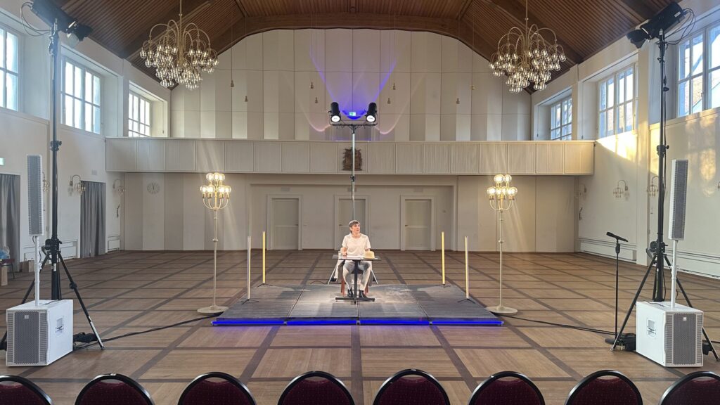 Mario Fuchs sitzt an einem Tisch auf einer Bühne und liest aus einem Buch vor