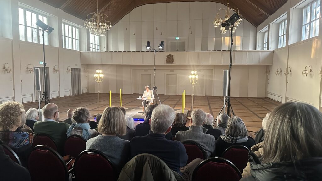 Mario Fuchs sitzt an einem Tisch auf einer Bühne und liest aus einem Buch vor