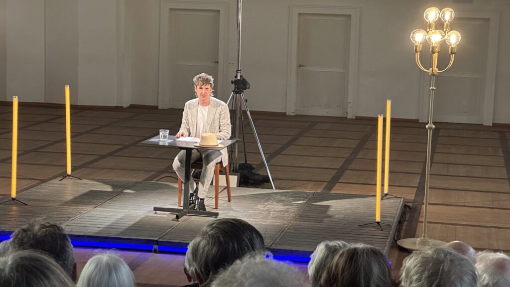 Mario Fuchs sitzt an einem Tisch auf einer Bühne und liest aus einem Buch vor