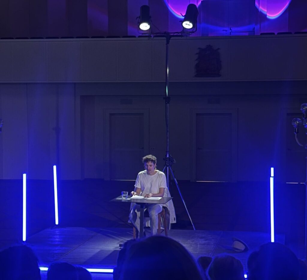 Mario Fuchs sitzt an einem Tisch auf einer Bühne und liest aus einem Buch vor