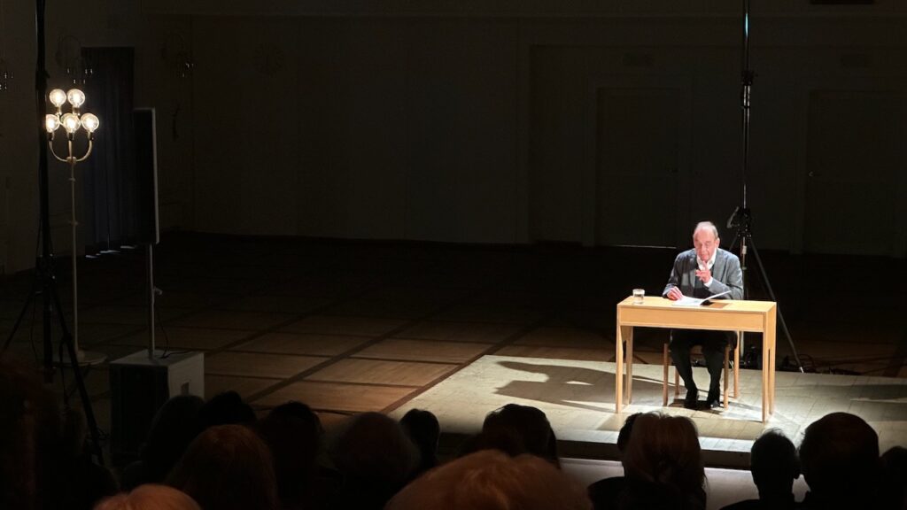 Peter Schröder sitzt an einem Tisch auf einer kleinen Bühne und liest vor.