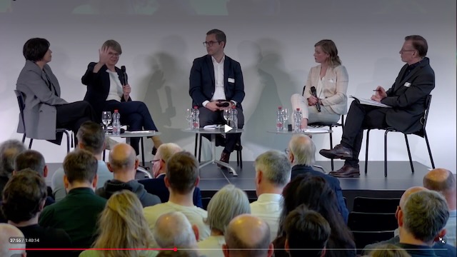 Podiumsdiskussion mit mehreren Teilnehmenden. Im Vordergrund ist das Publikum von hinten sichtbar.